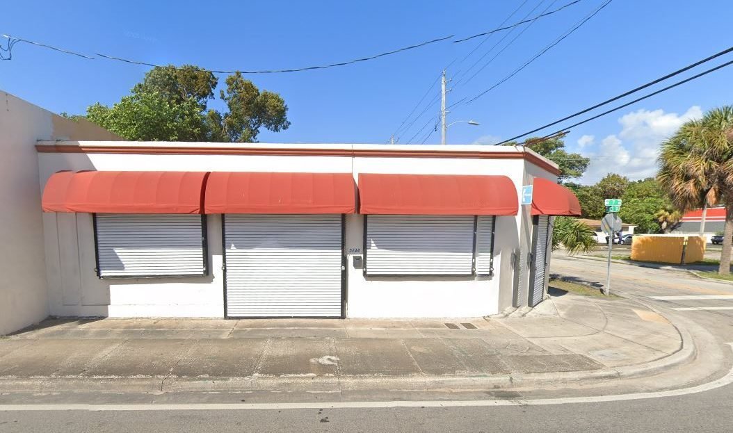 Retail space available for sale in Miami, FL. It is a one story white building with red awnings pictures on a day with blue skies.